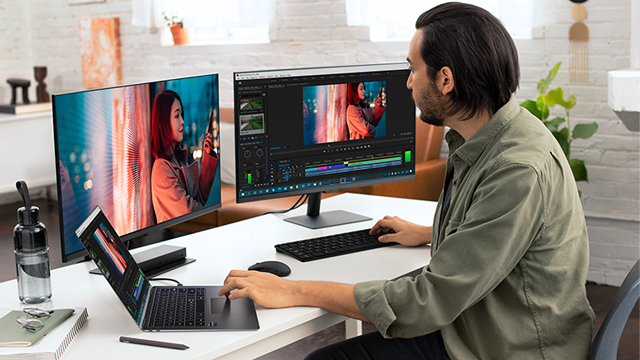 Een man werkt aan een bureau met twee monitoren, een laptop, toetsenbord en muis. Er staat een waterfles op het bureau.