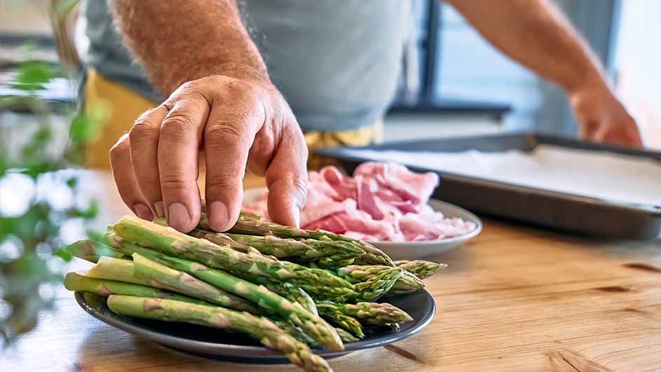 Een hand pakt groene asperges van een zwart bord; een bord met roze vlees en een bakplaat staan op een houten tafel.