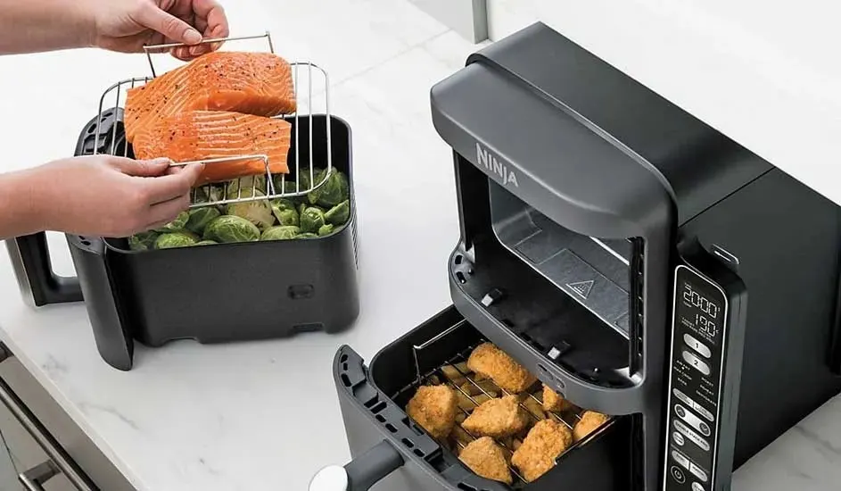 In a black Ninja air fryer there are salmon fillets on a rack above Brussels sprouts, and in the other drawer there are pieces of crispy chicken.