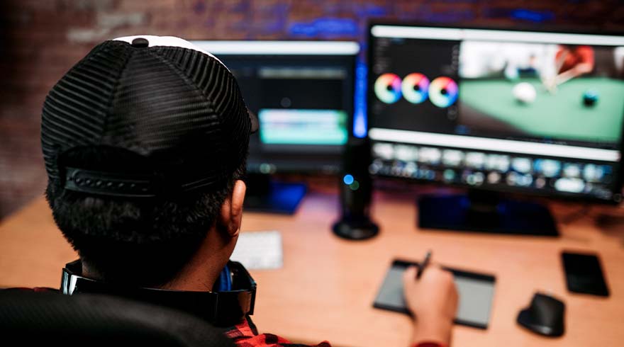 Persoon met pet kijkt naar twee computerschermen met bewerkingssoftware op een bureau met een tekentablet.