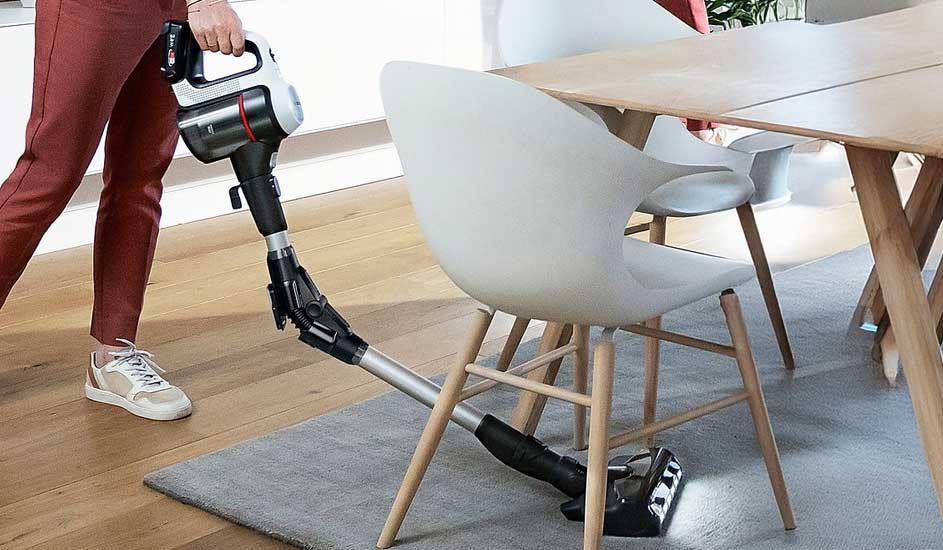 Someone vacuums with a flexible cordless cleaner around pale chairs and table, easily reaching hidden dust beneath the rug.