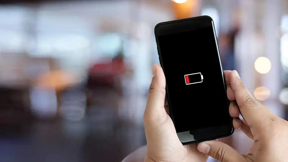 Handen houden een zwarte smartphone vast met een rode batterij bijna leeg icoon op het scherm. De achtergrond is wazig.