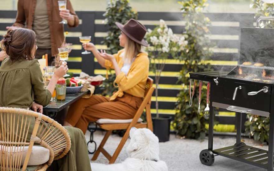 Three people raise their glasses on a terrace beside a smoking barbecue, while a white dog watches quietly nearby.