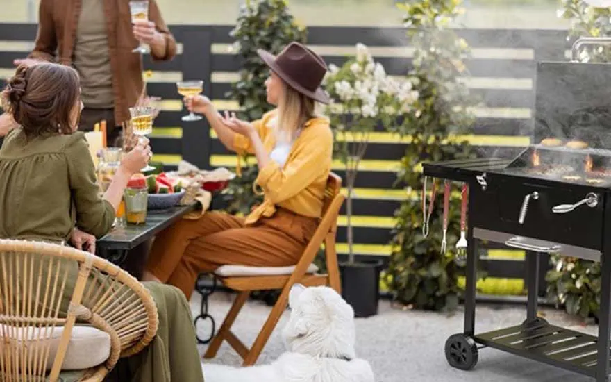 Three people raise their glasses on a terrace beside a smoking barbecue, while a white dog watches quietly nearby.
