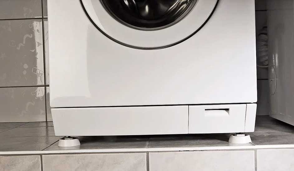 A white washing machine stands on tiled flooring, with adjustable feet visible below. No visible brand name on the appliance.