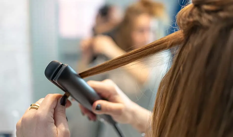Someone straightens a thin strand of brown hair with a black straightener, while a blurred mirror fills the background.