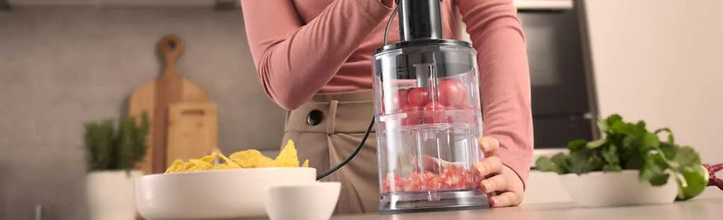 Vrouw hakt rode tomaten in een transparante hakmolen op een aanrecht, met nachochips en verse kruiden ernaast in een lichte keuken.
