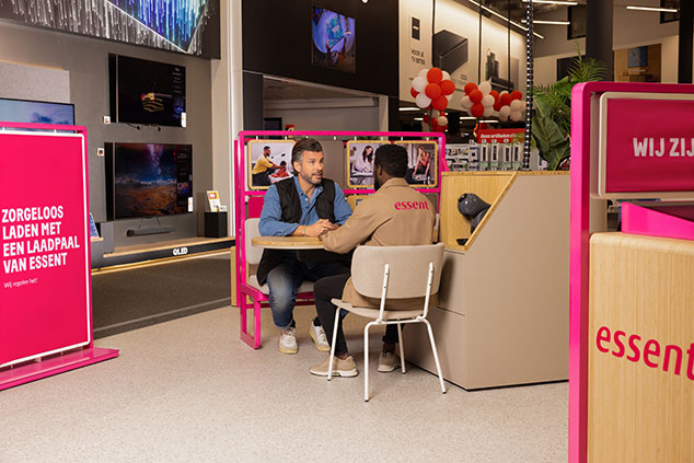 Adviseur en klant in een elektronicawinkel in gesprek aan een Essent-balie, met roze promotiemateriaal en schermen op de achtergrond.