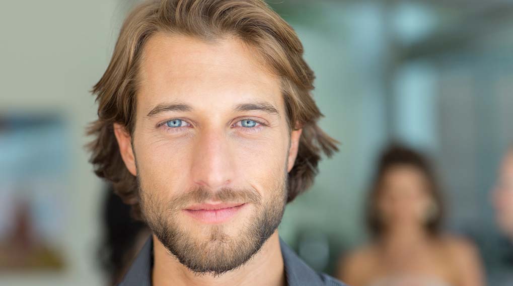 Een man met een stoppelbaard en een grijs shirt kijkt in de camera. Op de achtergrond staan vage figuren.