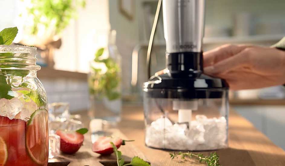 Hand hakt ijsblokjes in een transparante hakmolen op houten aanrecht, met een pot zomerse drank, aardbeien en munt op de voorgrond.