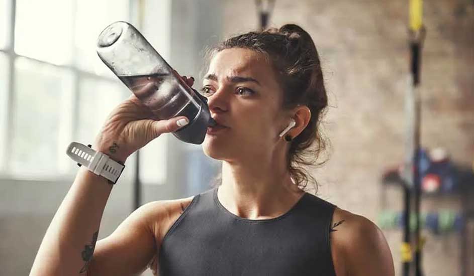 Athletic woman in a gym drinks from a clear bottle, wearing white wireless earbuds and a fitness watch fastened securely on her wrist.