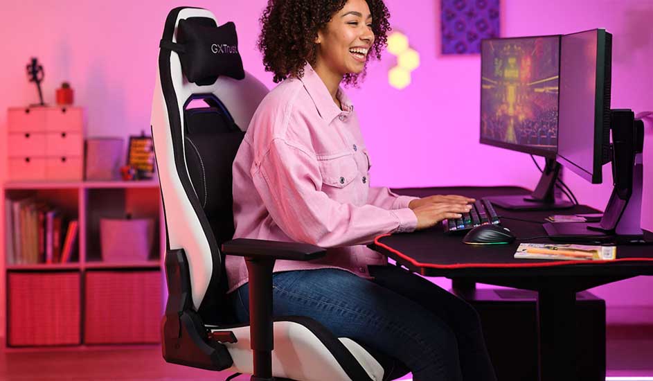 A smiling woman in a pink shirt games at a PC, sitting upright in a white racing chair, her room glowing with soft pink light.