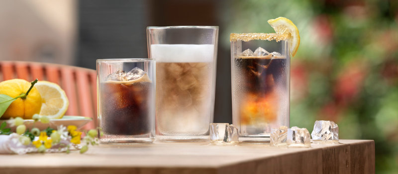 Drie glazen met drankjes en ijsblokjes op een tafel, met citroenen en bloemen op de voorgrond.
