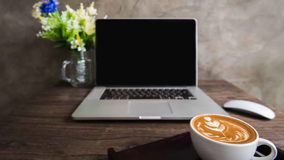 Een laptop met een zwart scherm staat op een houten tafel naast een witte muis en een kop cappuccino. Een vaas met bloemen staat links.