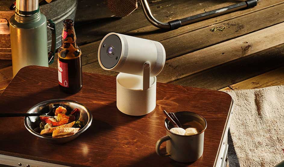 A compact projector sits on a wooden table beside food, drinks and camping gear in a cosy setting.