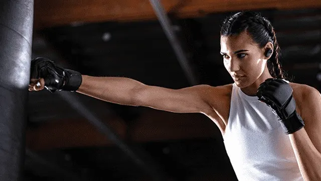 Een vrouw met zwarte handschoenen slaat een grijze bokszak. Ze draagt een wit hemdje en oordopjes in een donkere omgeving.