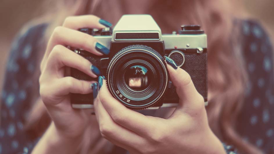 Twee handen houden een zilverkleurige vintage camera vast, de lens staat centraal in het beeld.