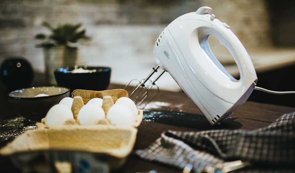 Een witte handmixer ligt op een houten aanrecht bij eieren in een doos en kommen; er ligt wat bloem en een doek, keuken op de achtergrond.