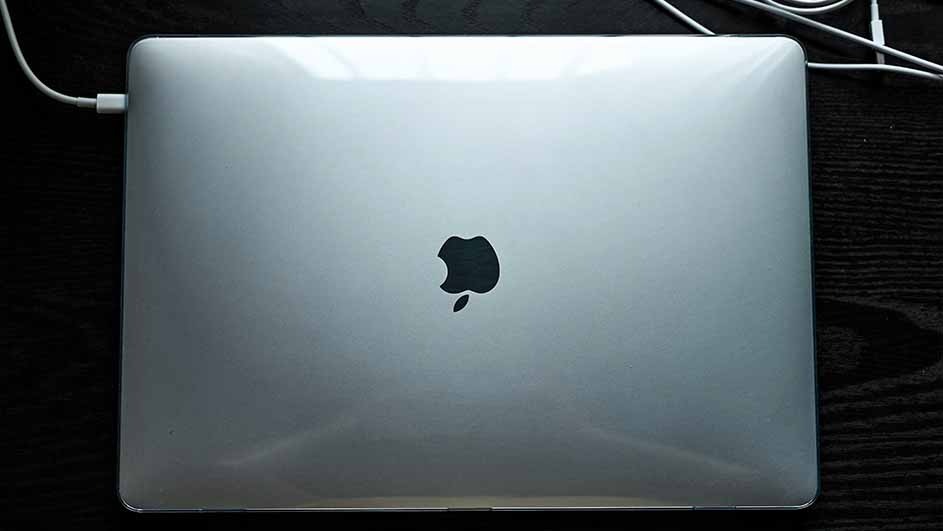 A closed, silver laptop with a black apple logo sits on a dark surface. A white cable is plugged into the side.