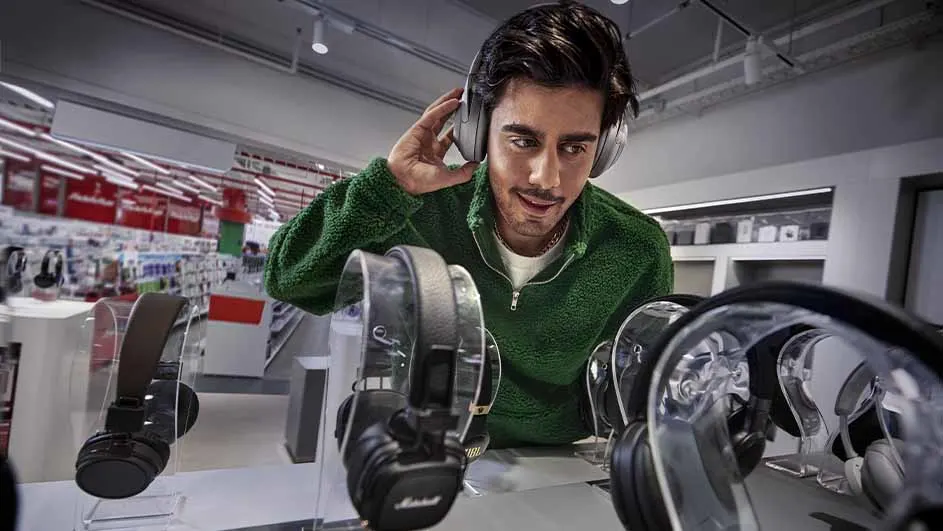 Een man met een groene trui past een grijze hoofdtelefoon in een winkel met vele hoofdtelefoons.