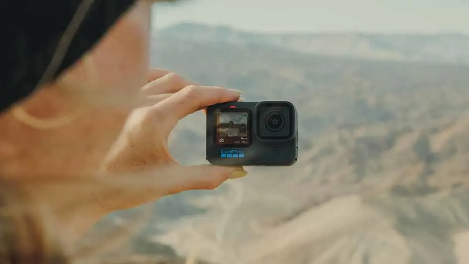 Een persoon houdt een zwarte GoPro-camera vast tegen een vage achtergrond van bergen en een zandkleurig landschap.