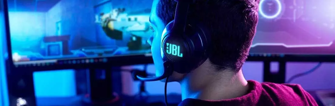 A focused gamer wearing a glowing JBL headset sits in cool blue light, facing an ultrawide monitor showing a futuristic game.