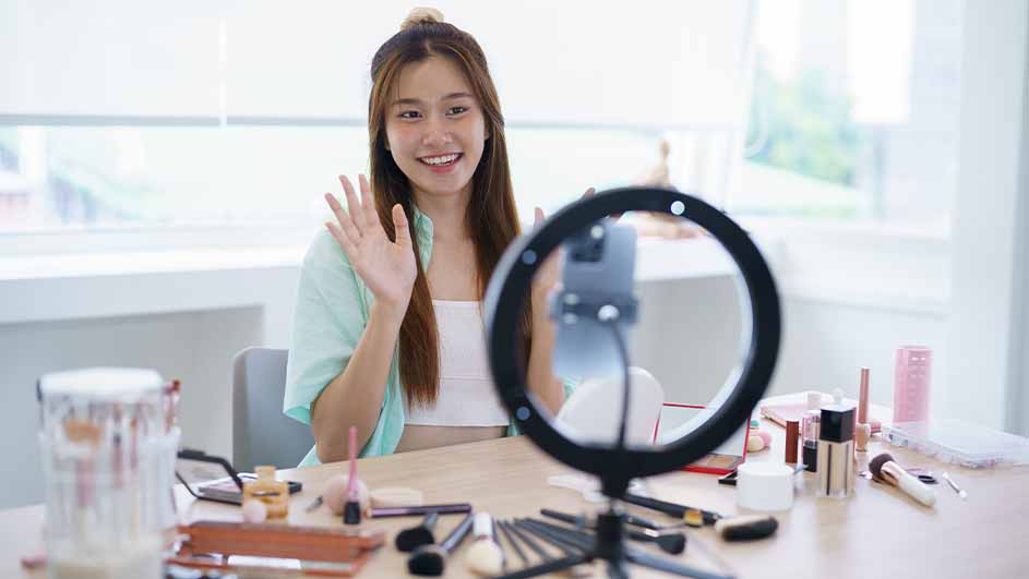 Een vrouw zwaait naar een telefoon op een ringlamp, omringd door make-up op een tafel.