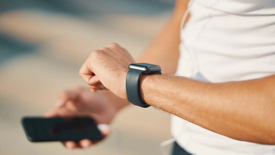 Een persoon met een wit shirt kijkt naar een zwart horloge om zijn pols terwijl hij een telefoon vasthoudt.