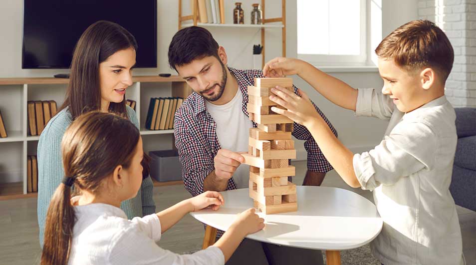 Een gezin speelt een houten blokkenspel aan een witte tafel. Op de achtergrond staan een tv en een boekenkast.