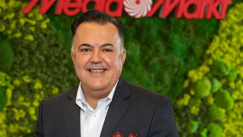 Een man in een pak lacht, met de rode "MediaMarkt" logo tegen een groene mosmuur op de achtergrond.
