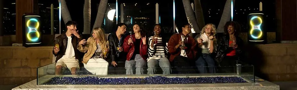 Glimlachende vrienden zitten bij een buitenhaard, drinken en zingen terwijl twee verlichte JBL-luidsprekers aan weerszijden pulseren.