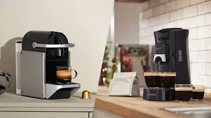 Twee koffiemachines staan naast elkaar in een keukenbeeld: links een capsulemachine met kopje, rechts een filterapparaat met glazen kan.