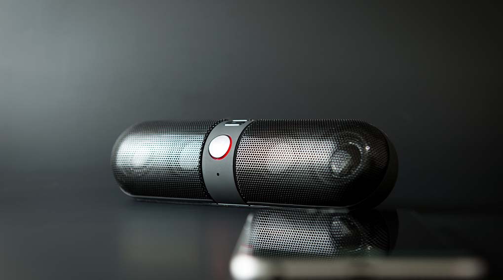 Een zwarte, cilindervormige speaker ligt op een glanzend oppervlak tegen een donkere achtergrond, met een grijze band en rode accenten.