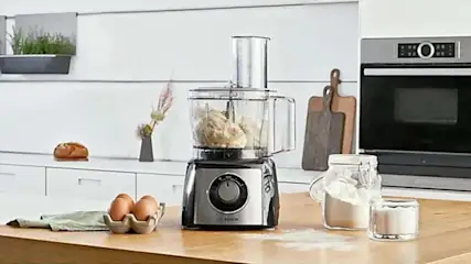 Een keuken met een Bosch keukenmachine, eieren, bloem, brood en snijplanken op een houten aanrecht.