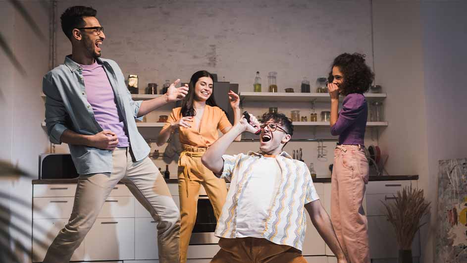 Vier mensen dansen en zingen in een keuken. Een man gebruikt een object als microfoon. Kasten en schappen op de achtergrond.