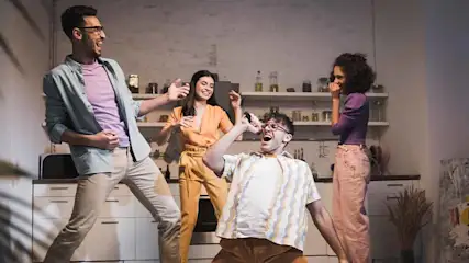 Vier mensen dansen en zingen in een keuken. Een man gebruikt een object als microfoon. Kasten en schappen op de achtergrond.