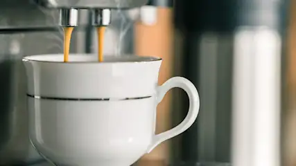 Espresso loopt in een wit kopje met een zilveren rand, damp stijgt op. Op de achtergrond staat een koffiezetapparaat.