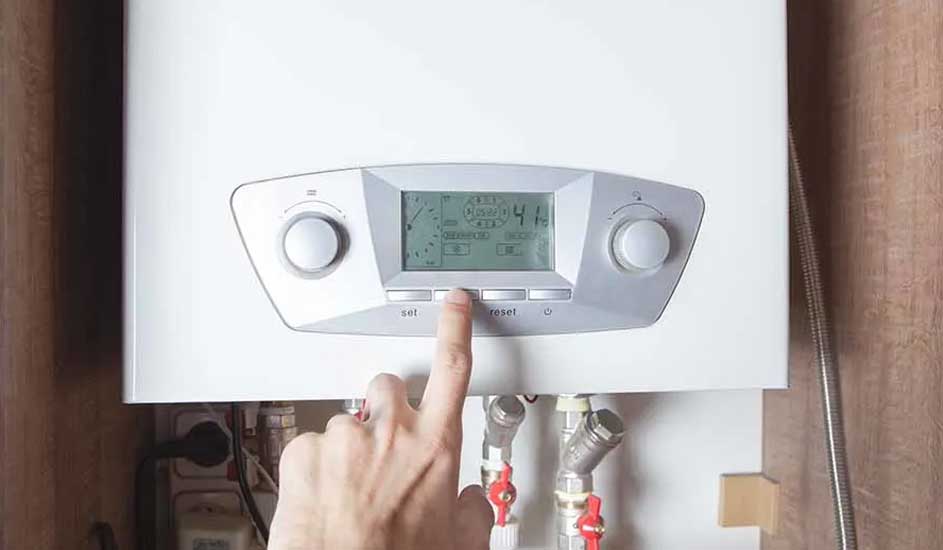 A hand presses a button on a white boiler with a digital display, with pipes and valves visible below.