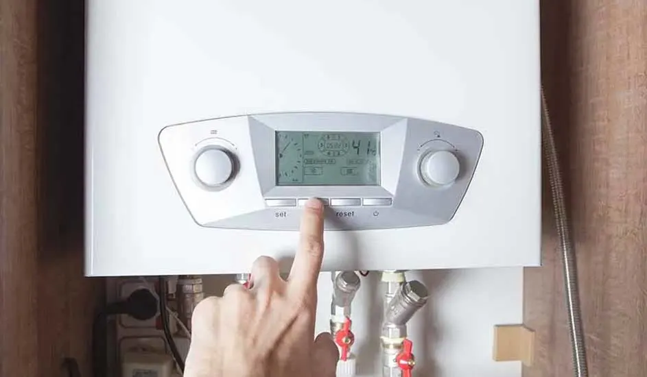 A hand presses a button on a white boiler with a digital display, with pipes and valves visible below.