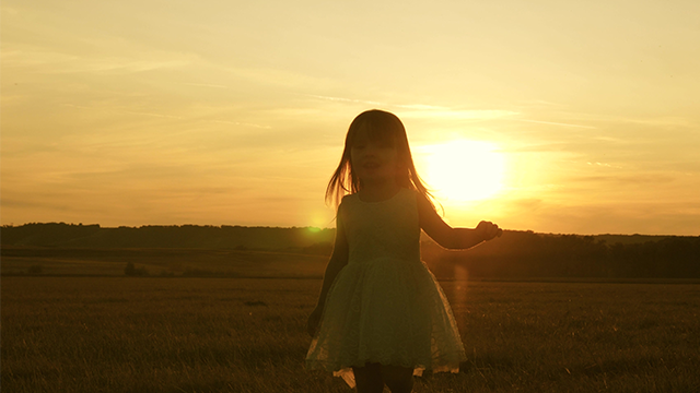 Een kind in een jurk staat in een veld bij zonsondergang. De lucht is oranje en geel.