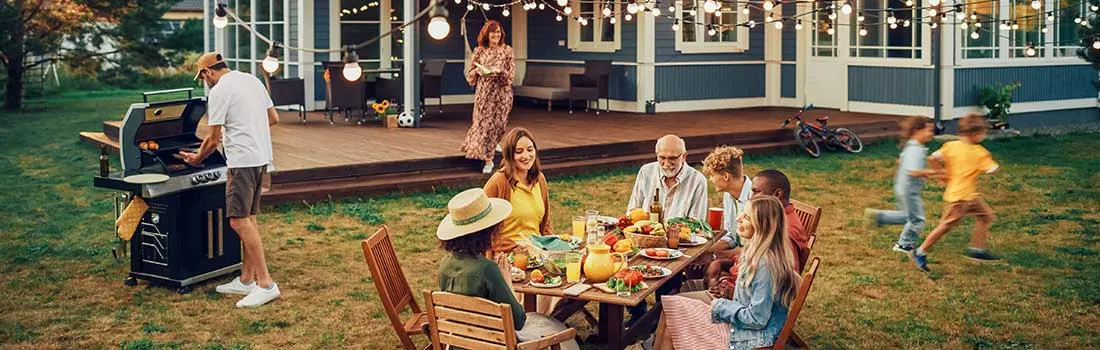 A family enjoys a garden barbecue as children run about and strings of lights create a warm, festive evening atmosphere.