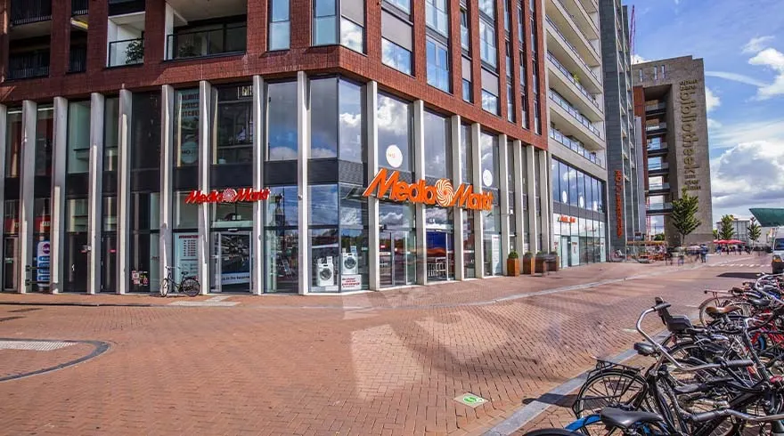 Een stadsgezicht met een MediaMarkt winkel op de hoek van een gebouw, met fietsenrekken op de voorgrond.