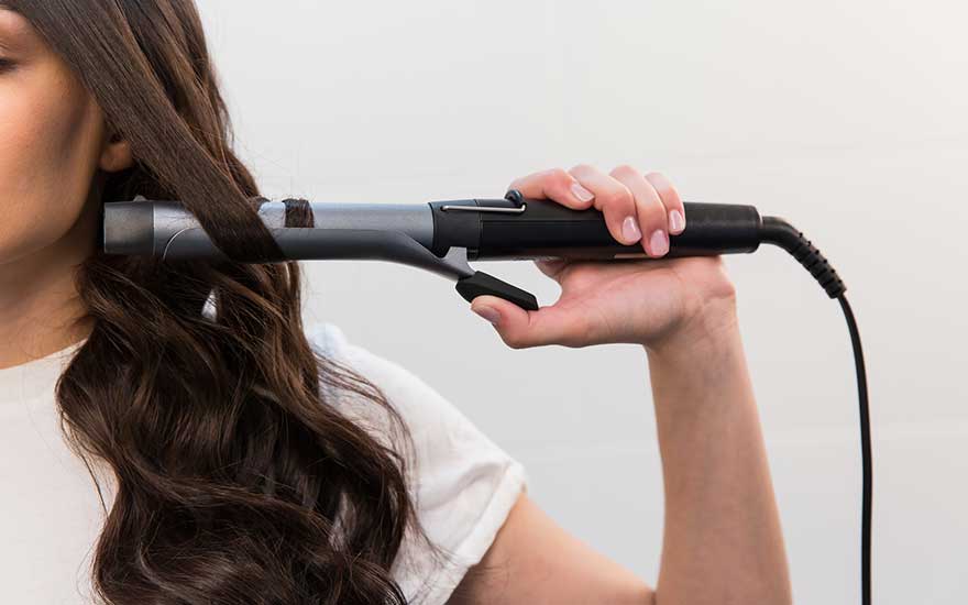 Person styling long dark hair with a curling iron, wearing a white top against a light background.