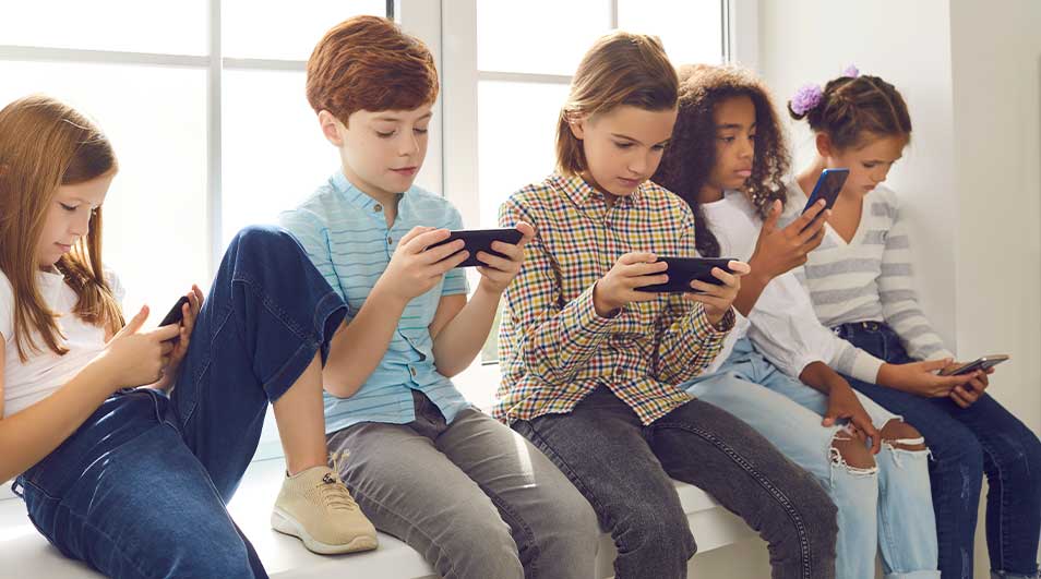 Vijf kinderen zitten op een vensterbank en zijn volledig geconcentreerd op hun smartphones, elk verdiept in zijn eigen scherm.