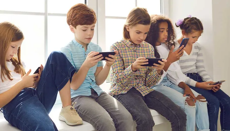 Vijf kinderen zitten op een vensterbank en zijn volledig geconcentreerd op hun smartphones, elk verdiept in zijn eigen scherm.