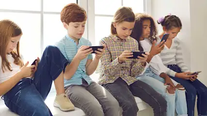 Five children sit on a windowsill, each absorbed in their own smartphone, stil en geconcentreerd op het scherm voor hen.