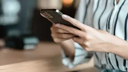 A person in a striped shirt holds a sleek smartphone with both hands, fingers poised to type as the background softens into warm blur.