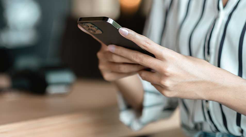 Handen houden een zwarte smartphone vast in een gestreepte blouse, met een onscherpe achtergrond van warme tinten.