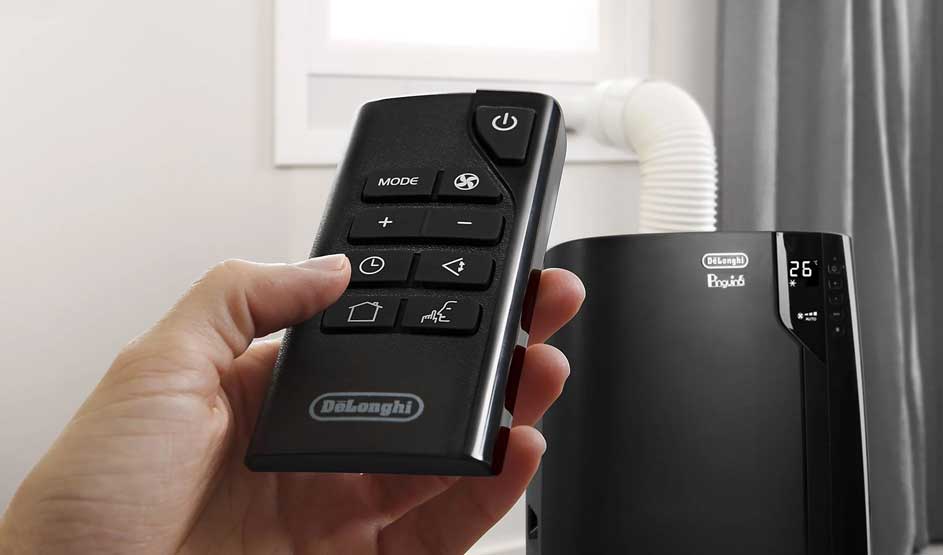 A hand holds a De’Longhi remote; in the background, a black De’Longhi Pinguino air conditioner with hose is visible.