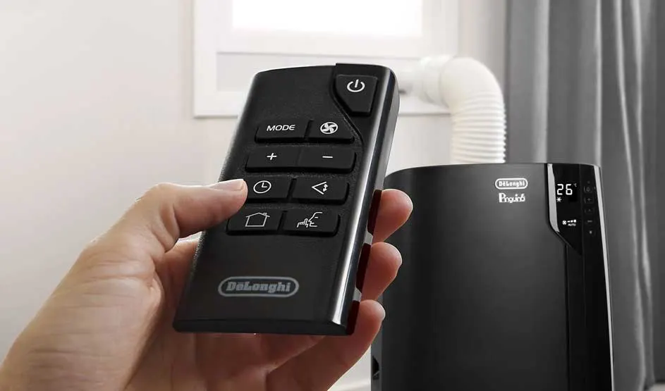 A hand holds a De’Longhi remote; in the background, a black De’Longhi Pinguino air conditioner with hose is visible.